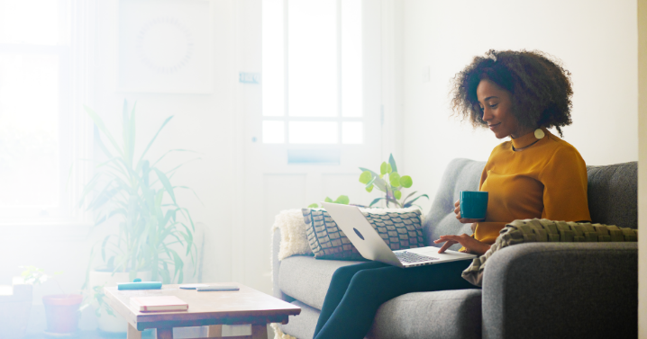 A woman is working on her laptop. She is sitting on a sofa and holding a mug.