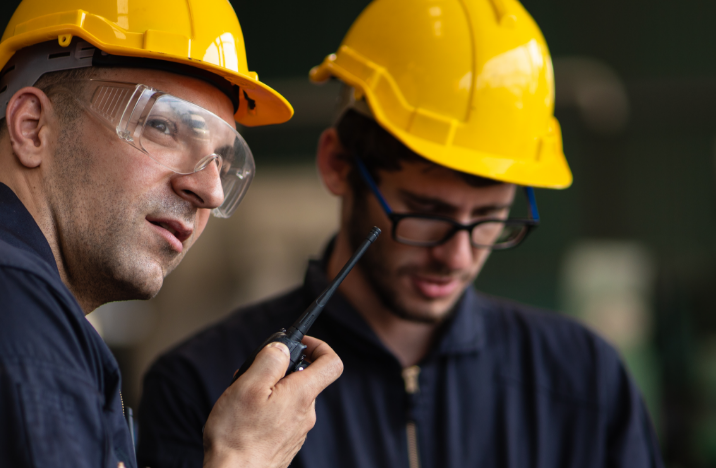 Two construction workers are standing next to each other. One is looking to his left while the other is looking down.