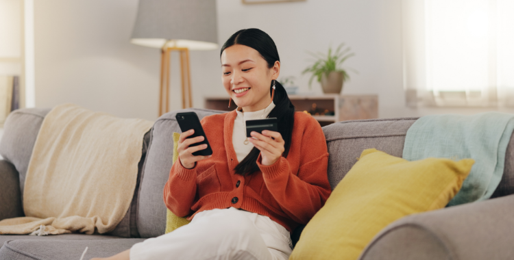 A woman is sitting on a sofa with a credit card in one hand, looking excitedly at the phone in her other hand.