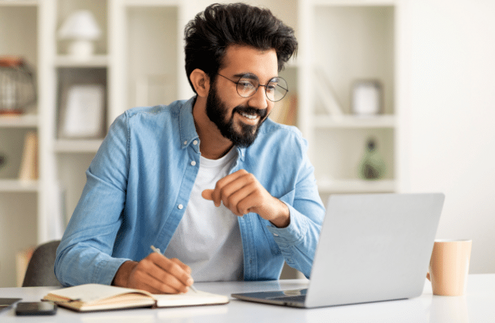 A man is working on his laptop while writing in a notebook.