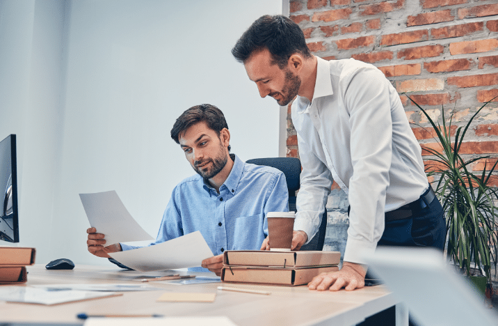 Two men are discussing a paper. One is sitting while the other is standing next to him.